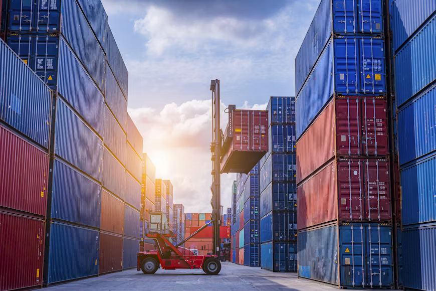 Stacked shipping containers in a logistics yard, with a reach stacker moving a container between the rows.